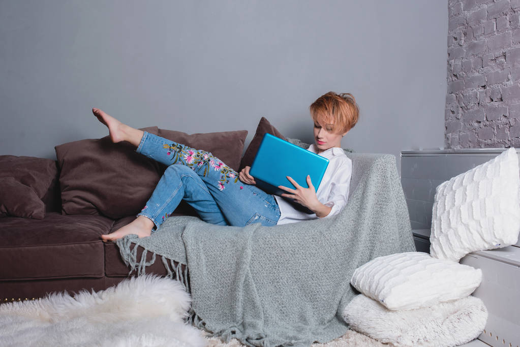 Stijlvolle jonge vrouw met laptop in handen op de sofa en laptop kijken. Gekleed in een wit overhemd en jeans met borduursel. Grijze muur met schaduw achter haar. - Foto, afbeelding