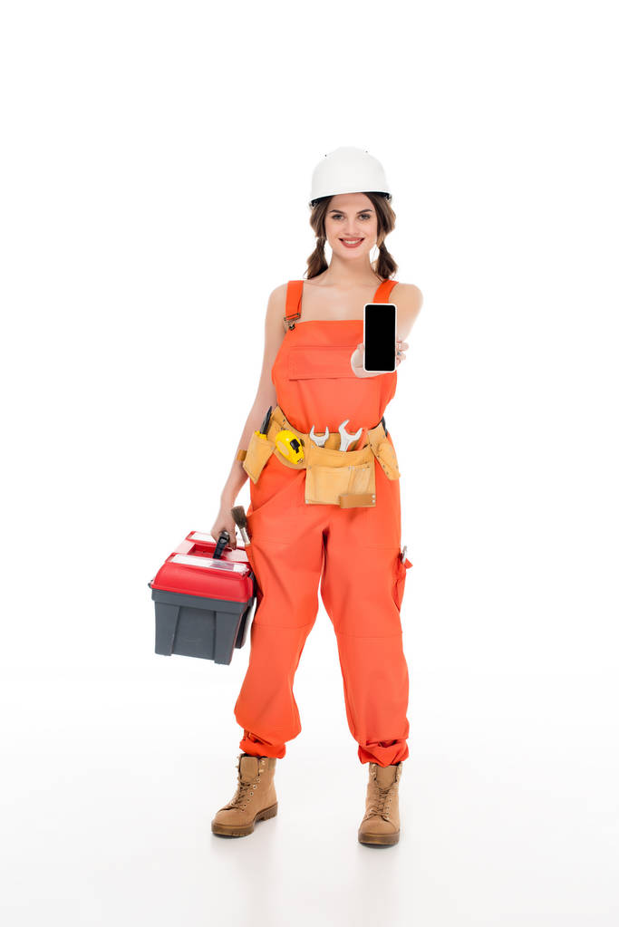 hermosa trabajadora sonriente en uniforme sosteniendo caja de herramientas y teléfono inteligente, aislado en blanco
 - Foto, imagen