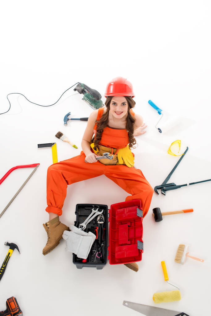 Overhead-Ansicht von Mädchen in Overalls auf dem Boden sitzend mit Werkzeugkiste und verschiedenen Geräten, isoliert auf weiß - Foto, Bild
