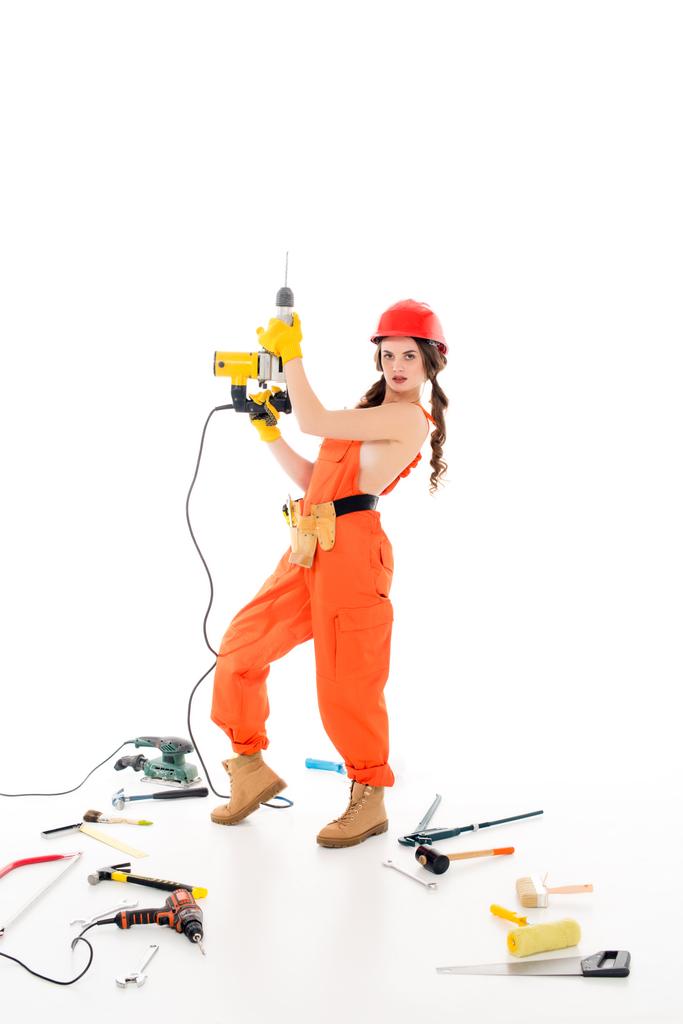 trabajadora en overol con taladros eléctricos, diferentes herramientas tumbadas en el suelo, aisladas en blanco
 - Foto, Imagen