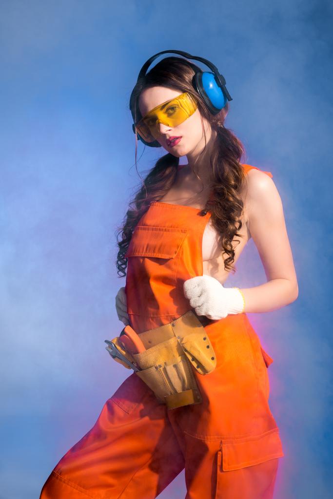 όμορφη σέξι κορίτσι με φόρμες με ζώνη εργαλείων, γυαλιά και προστατευτικά ακουστικά, απομονώνονται σε μπλε - Φωτογραφία, εικόνα