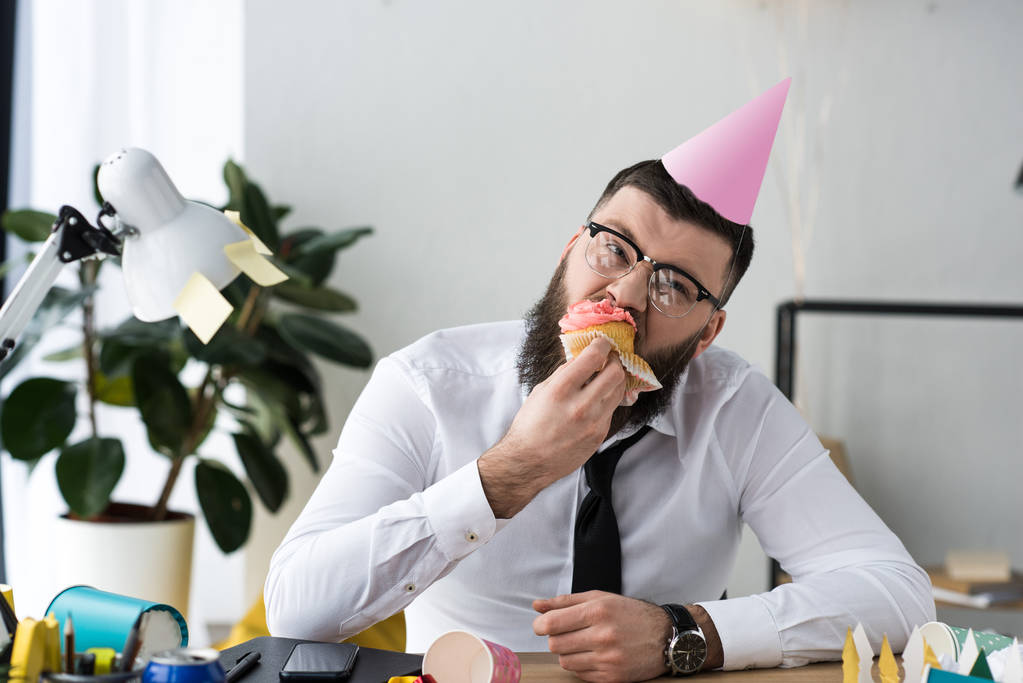 portrait of businessman in party cone eating birthday cupcake in office - Photo, Image