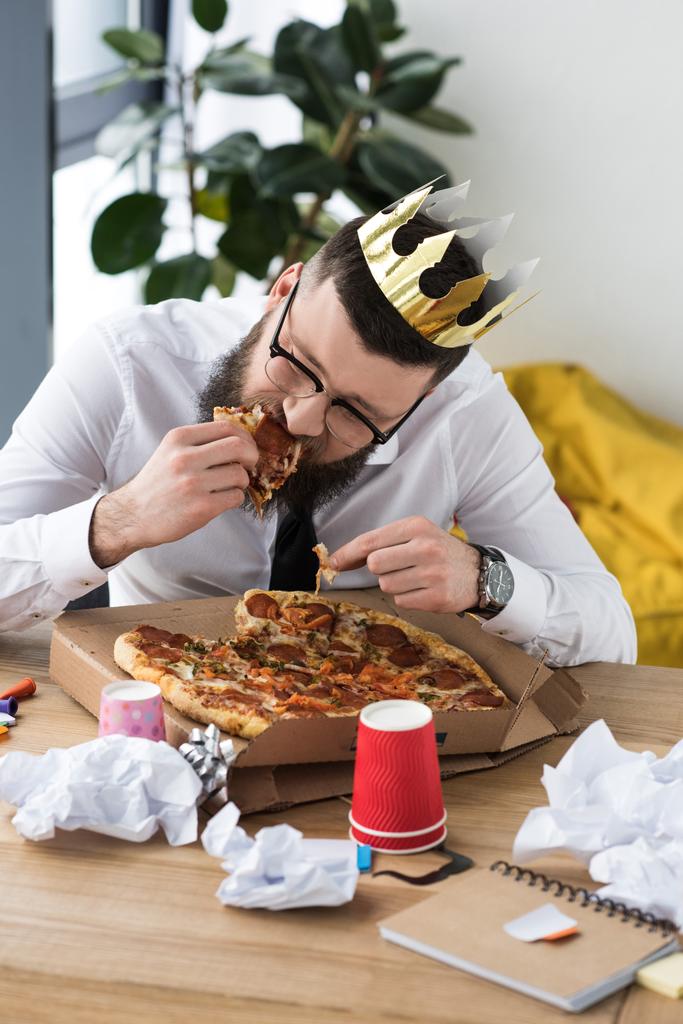 portrait of businessman with paper crown on head eating pizza at workplace - Photo, Image