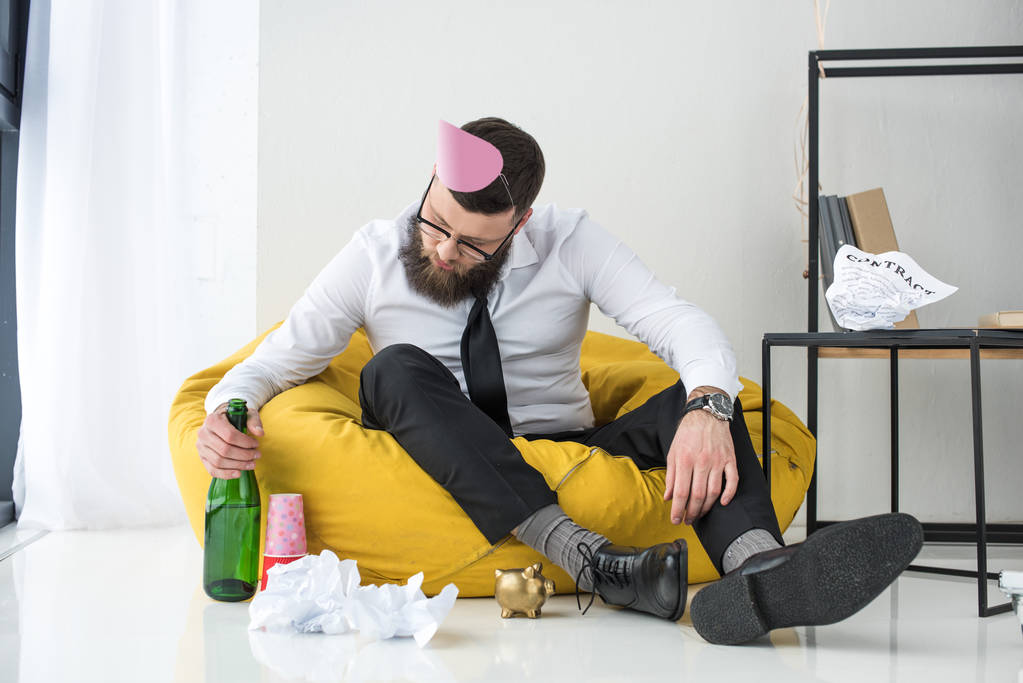 Empresario borracho en ropa formal con cono de papel en la cabeza sentado en la silla del bolso
 - Foto, imagen