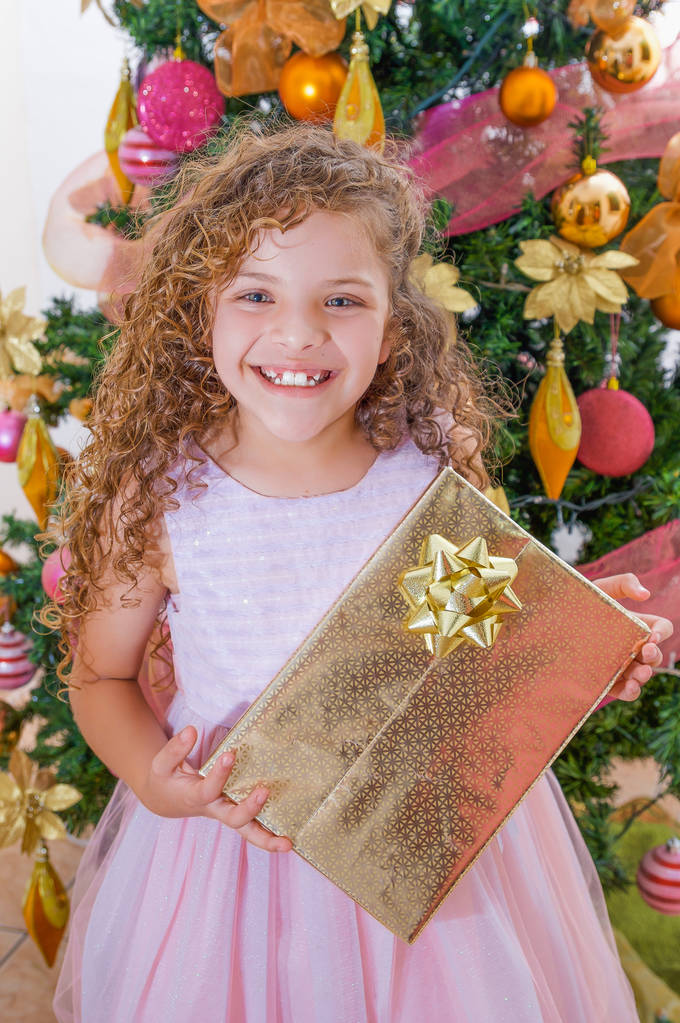 Bella ragazza riccia sorridente indossa un abito rosa e tenendo in mano un enorme regalo, con un albero di Natale dietro, Natale e concetto di nuovo anno
 - Foto, immagini