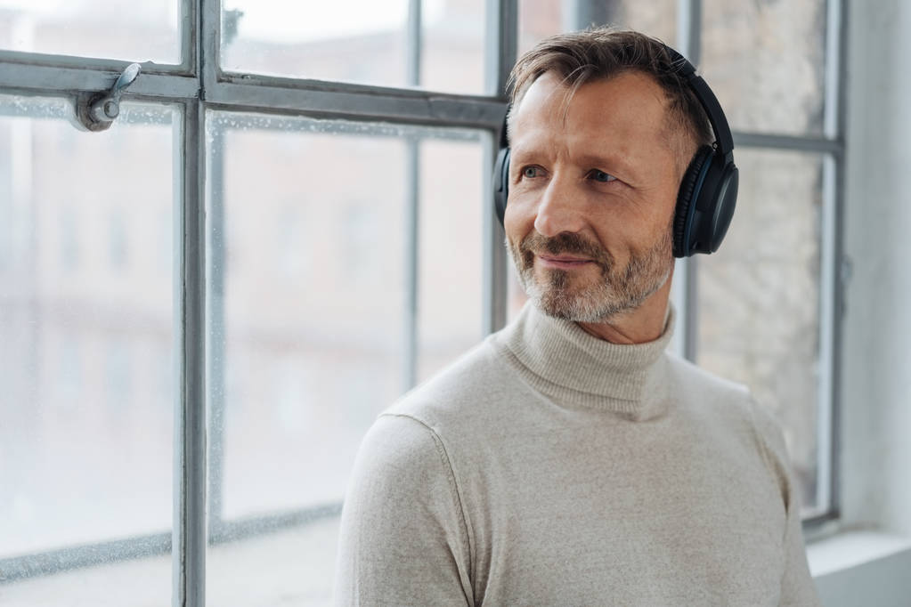 Усміхнений чоловік середнього віку слухає свою музику, коли він спирається на підвіконня, що повертається, щоб виглядати надворі
 - Фото, зображення