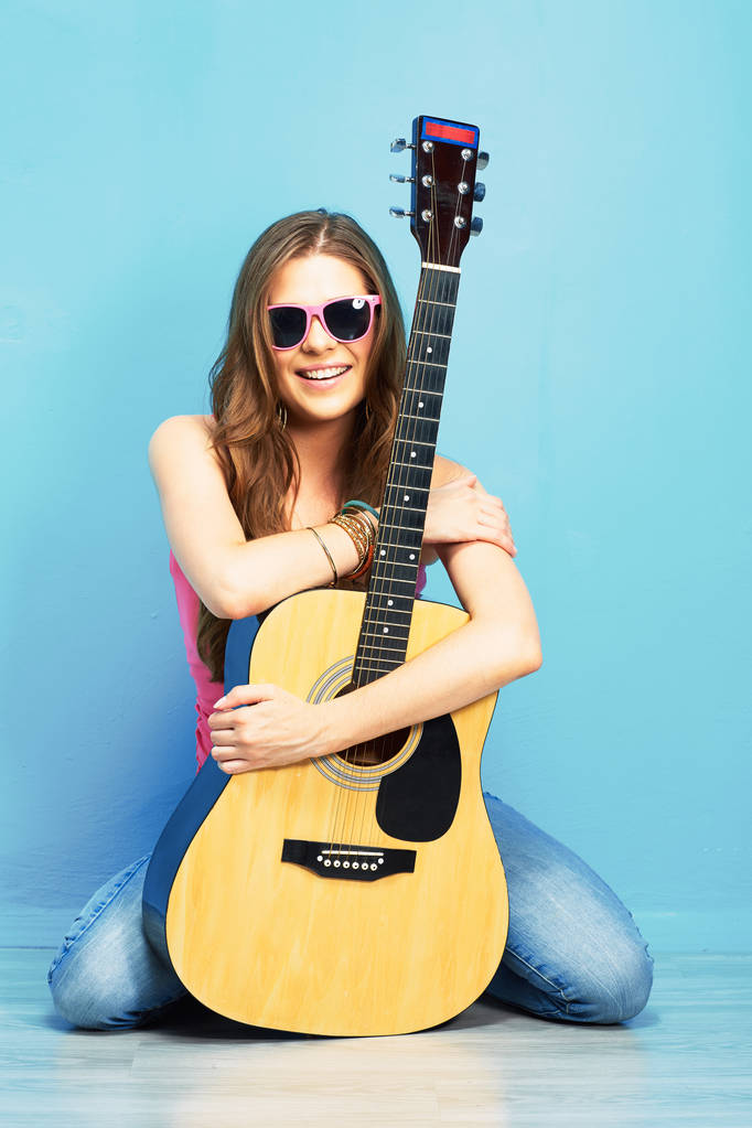 fashion music look with young model in sunglasses holding acoustic guitar and sitting on blue floor - Photo, Image
