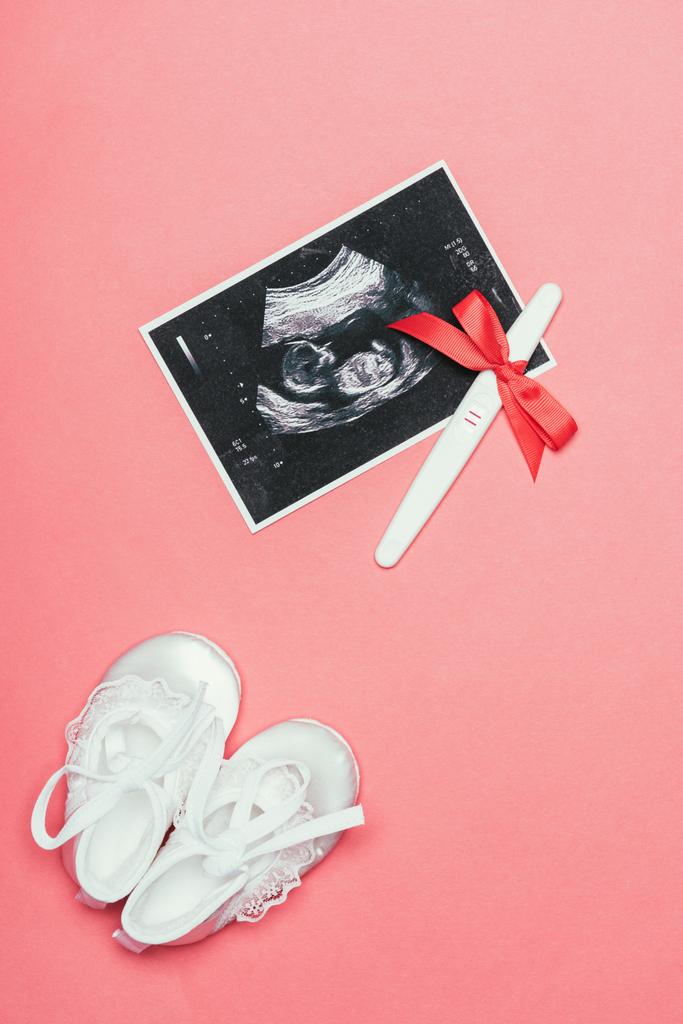 bovenaanzicht van plaatsing van kinderachtig schoenen, echografie en zwangerschapstest met lint geïsoleerd op roze - Foto, afbeelding