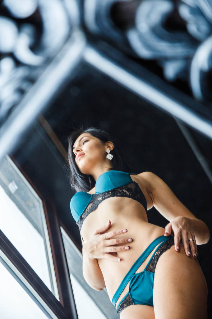 Студийный снимок красивой брюнетки в сексуальном нижнем белье. Девушка спортивного телосложения, стоящая у окна квартиры
 - Фото, изображение