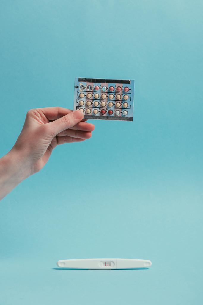 частичный взгляд женщины с противозачаточными таблетками в руке и тестом на беременность, изолированный на синем
 - Фото, изображение