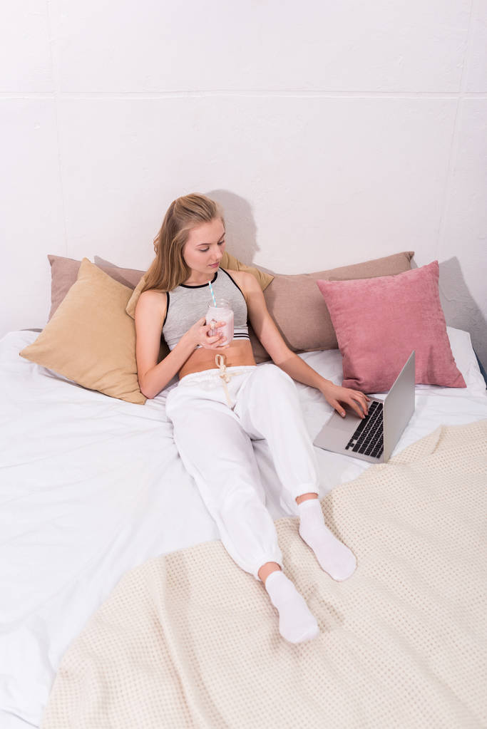 ベッドでノート パソコンを操作する石工の瓶にミルクセーキを持つ若い女性のハイアングル - 写真・画像