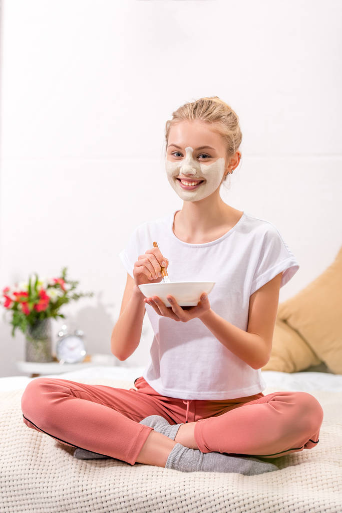 ευτυχισμένη γυναίκα εφαρμογή λευκό πηλό μάσκα στο πρόσωπο, ενώ κάθεται στο κρεβάτι - Φωτογραφία, εικόνα