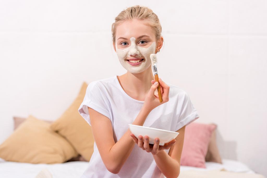 mujer joven aplicando máscara de arcilla blanca en la cara mientras está sentado en la cama
 - Foto, Imagen