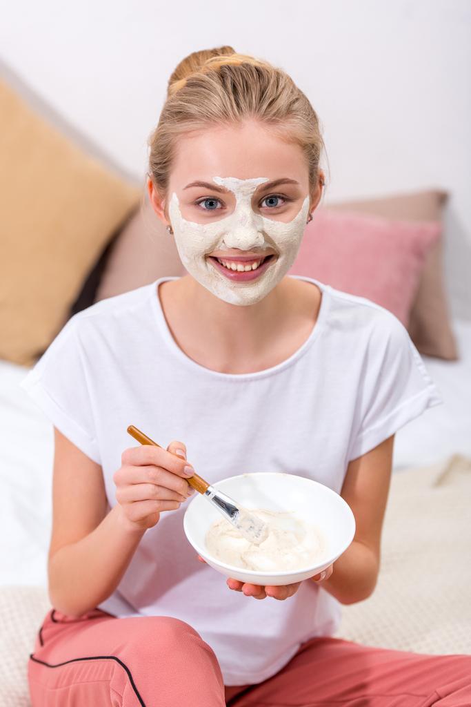 mujer joven aplicando máscara de arcilla en la cara en casa y mirando a la cámara
 - Foto, imagen