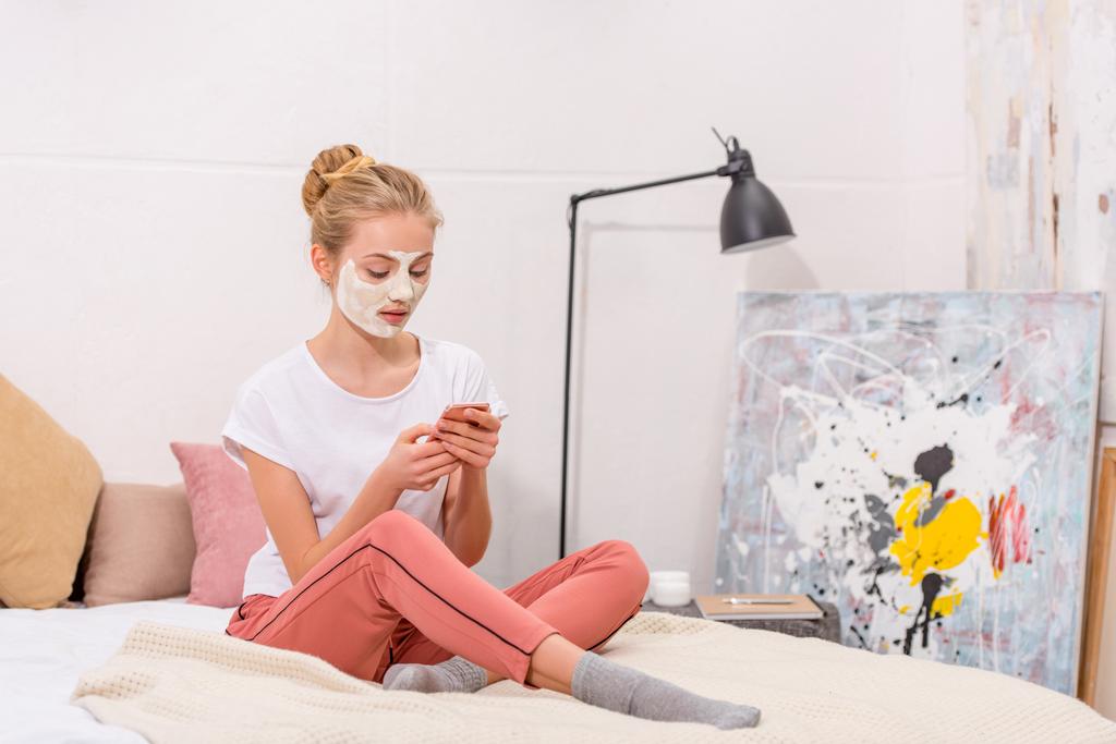 young woman with white clay facial mask using smartphone while sitting on bed at home - Photo, Image