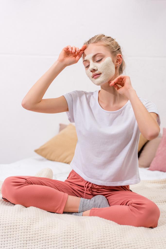 νεαρή γυναίκα με λευκό πηλό μάσκα προσώπου που κάθεται στο κρεβάτι στο σπίτι - Φωτογραφία, εικόνα