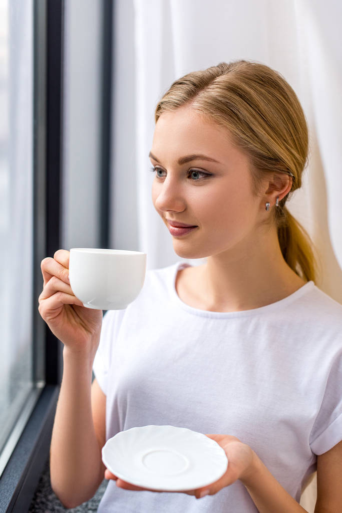 счастливая молодая женщина пьет кофе и смотрит в окно
 - Фото, изображение