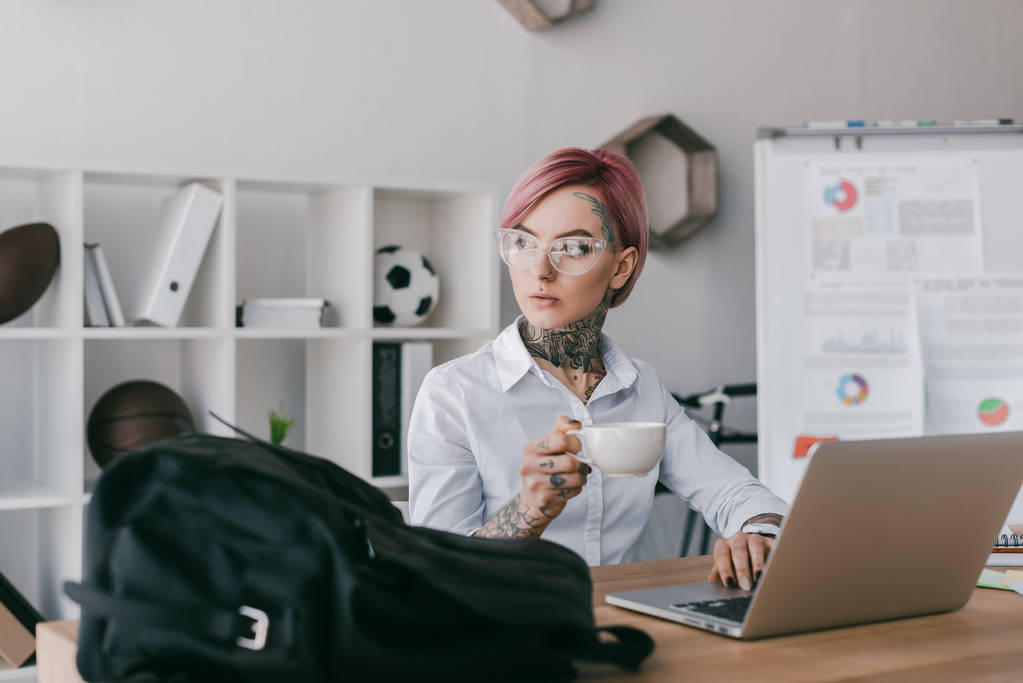 mladá podnikatelka držící šálek kávy a koukal při používání notebooku na pracovišti - Fotografie, Obrázek