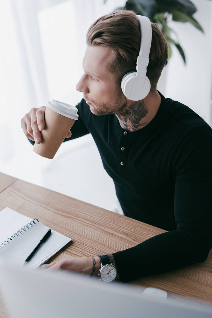 high angle view of young tattooed businessman in headphones drinking coffee from paper cup - Photo, Image
