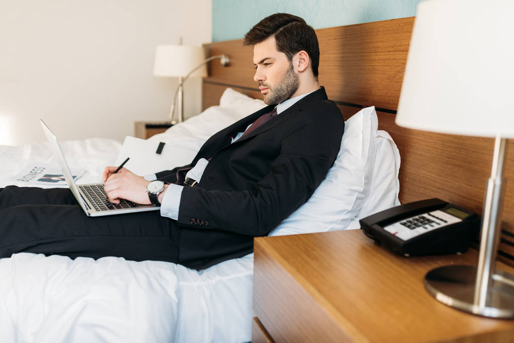 Μερική άποψη του επιχειρηματία ξαπλωμένος στο κρεβάτι σε δωμάτιο ξενοδοχείου και να χρησιμοποιούν φορητό υπολογιστή  - Φωτογραφία, εικόνα