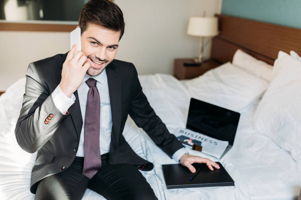 homme d'affaires souriant parlant par smartphone et assis sur le lit dans la chambre d'hôtel
 - Photo, image