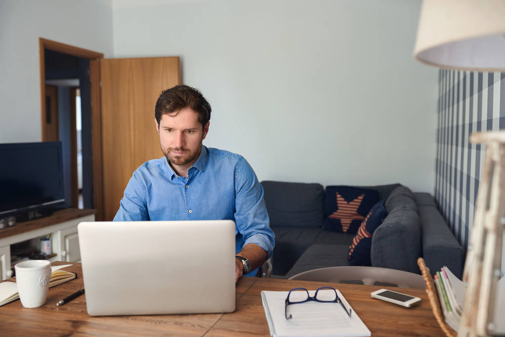 Mladý muž pracovat v režimu online s notebookem u stolu ve svém obývacím pokoji při práci sama doma - Fotografie, Obrázek