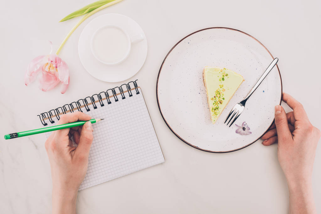 przycięte strzał robiąc notatki notebook i kawałek ciasta i filiżankę mleka w pobliżu przez kobiety - Zdjęcie, obraz