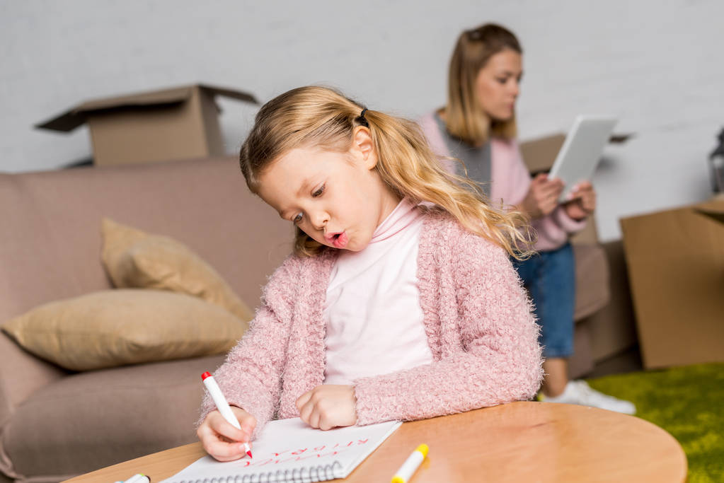 Kind zeichnet mit Filzstift, während Mutter digitales Tablet auf Sofa benutzt - Foto, Bild