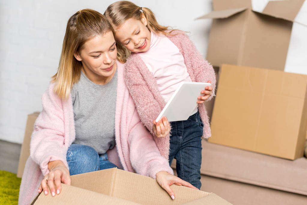 happy mother and daughter using digital tablet while packing cardboard boxes - Photo, Image