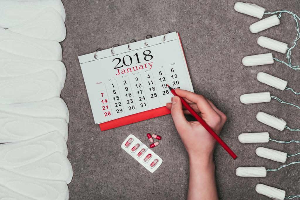 oříznutý snímek ženy ukazující na datum v kalendáři s prášky, menstruační vložky a tampony kolem na šedém povrchu - Fotografie, Obrázek