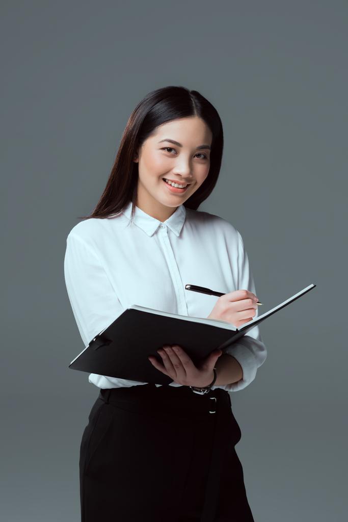 Lächelnde junge Geschäftsfrau macht sich Notizen und blickt vereinzelt in die Kamera - Foto, Bild