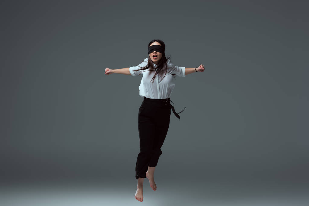 young barefoot woman in black blindfold jumping with open mouth on grey - Photo, Image