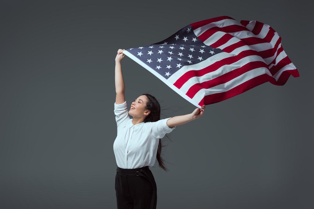 heureuse jeune femme tenant drapeau américain dans les mains levées et regardant loin isolé sur gris
 - Photo, image