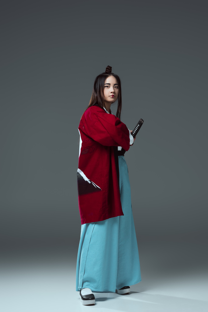 joven asiático mujer en kimono celebración katana espada y mirando a cámara en gris
 - Foto, Imagen