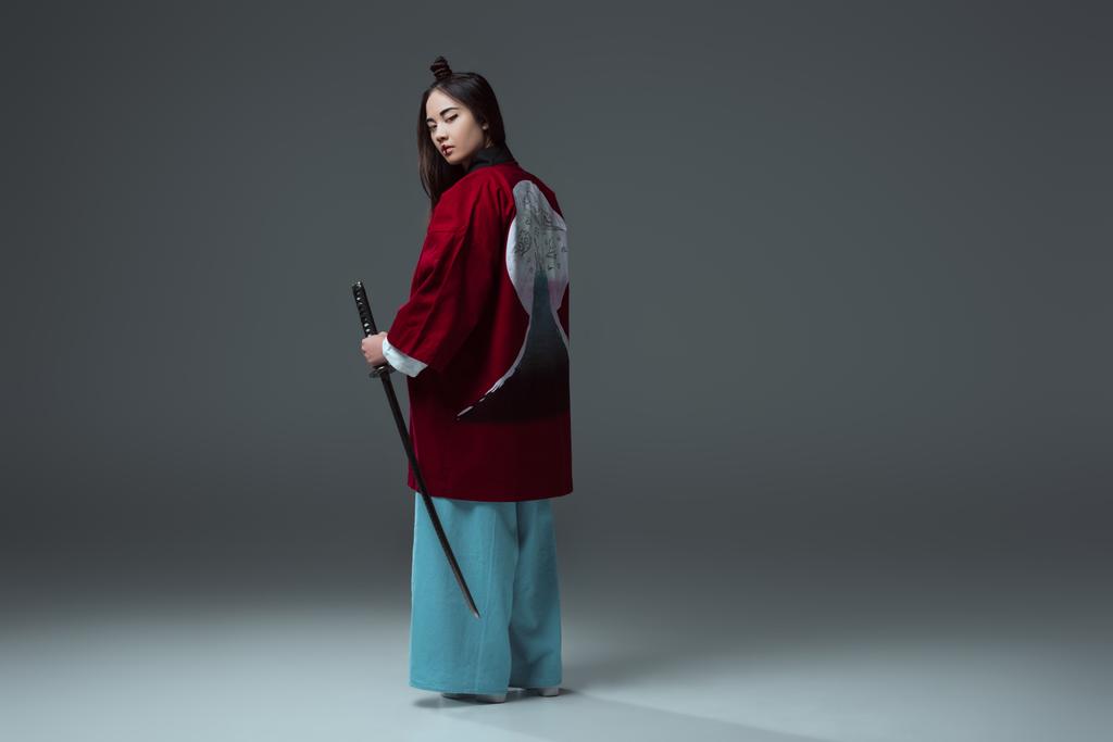 back view of samurai in kimono holding katana and looking at camera on grey   - Photo, Image