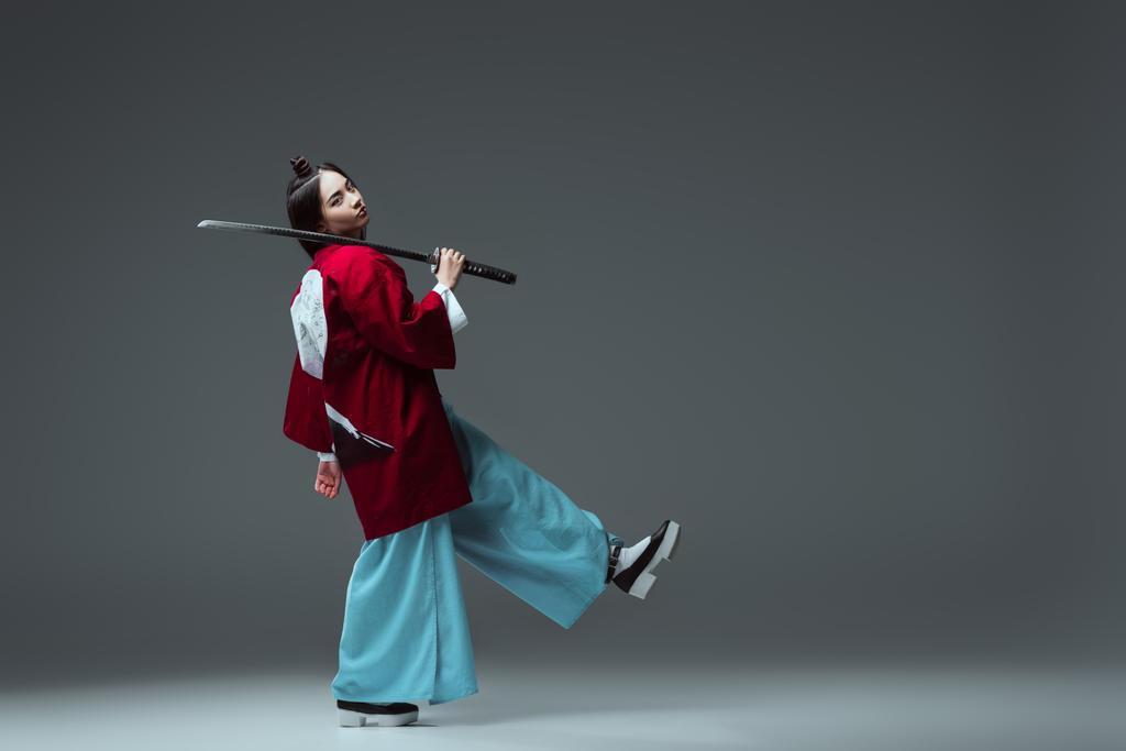 side view of samurai in kimono walking with katana and looking at camera on grey - Photo, Image