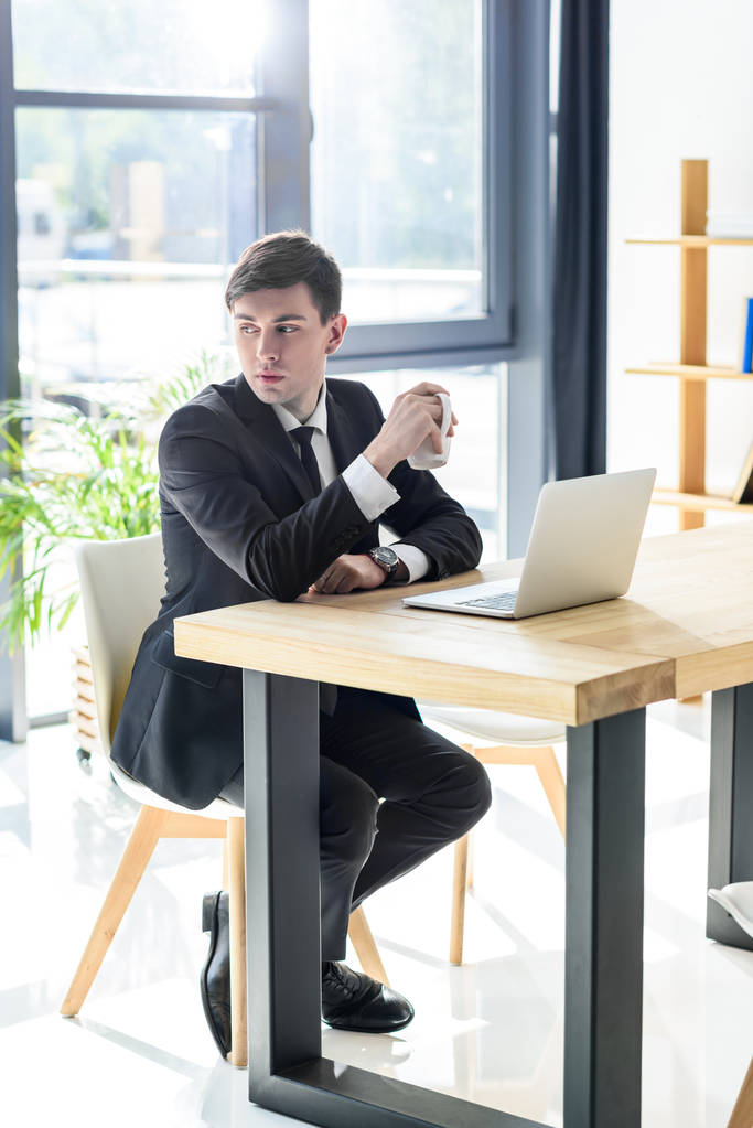 junger Geschäftsmann trinkt Kaffee, während er im modernen Büro am Laptop arbeitet   - Foto, Bild