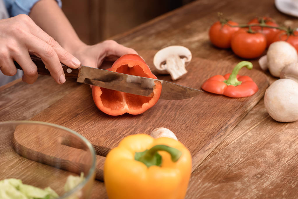 abgeschnittenes Bild einer Frau, die in der Küche rote Paprika auf einem Holzbrett schneidet - Foto, Bild