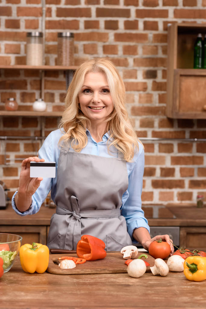 nainen osoittaa luottokortilla ja seisoo lähellä pöydän vihanneksia keittiössä
 - Valokuva, kuva