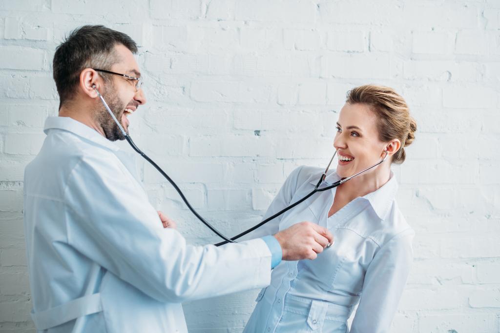 Lachende Ärzte hören einander mit Stethoskopen zu - Foto, Bild