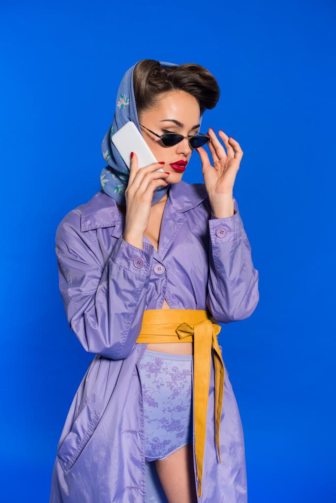 Portret van modieuze vrouw in retro-stijl kleding praten over smartphone geïsoleerd op blauw - Foto, afbeelding