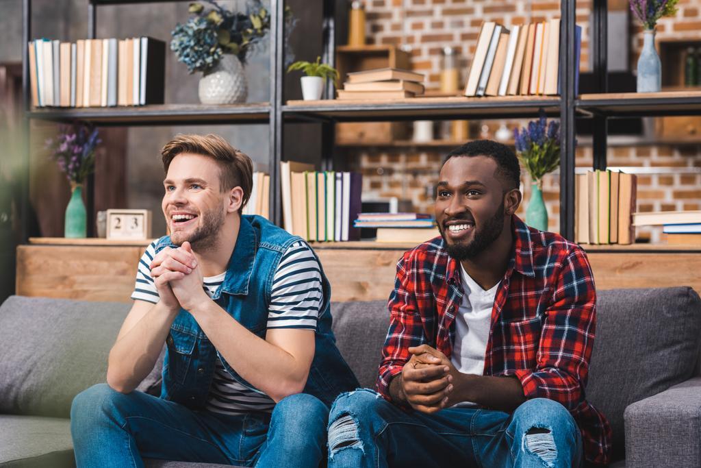 feliz joven multiétnico hombres sentados en el sofá y mirando hacia otro lado en casa
 - Foto, imagen
