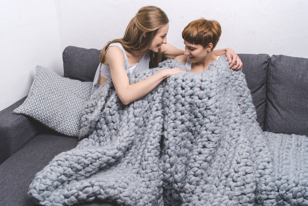 happy young lesbian couple under knitted wool blanket on couch - Photo, Image