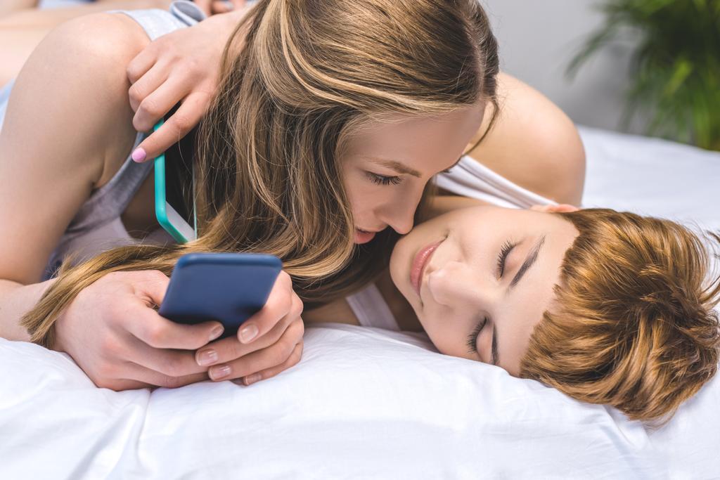 joven pareja lesbiana abrazándose en la cama mientras usa teléfonos inteligentes
 - Foto, imagen