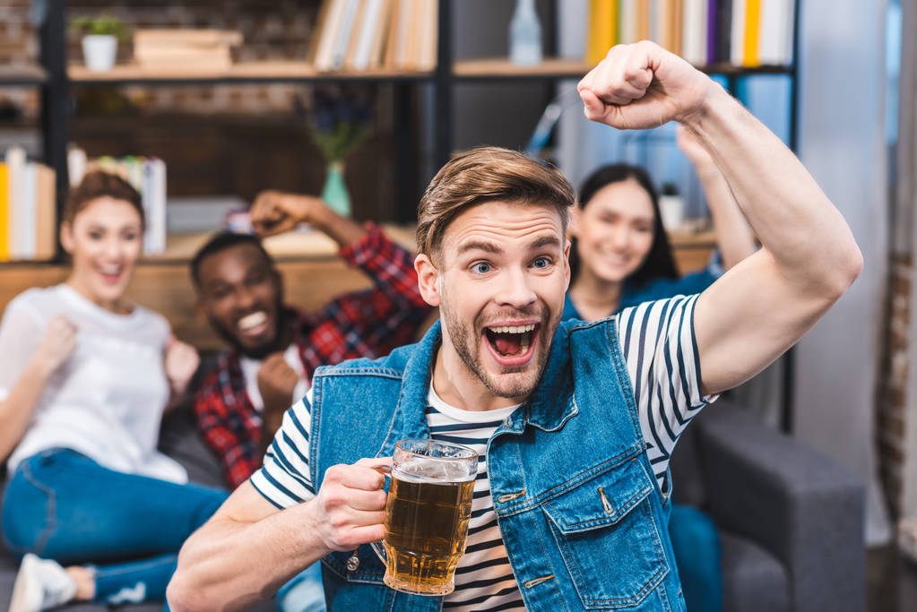 возбужденный молодой человек держит стакан пива и смотреть телевизор с друзьями сидя позади
 - Фото, изображение
