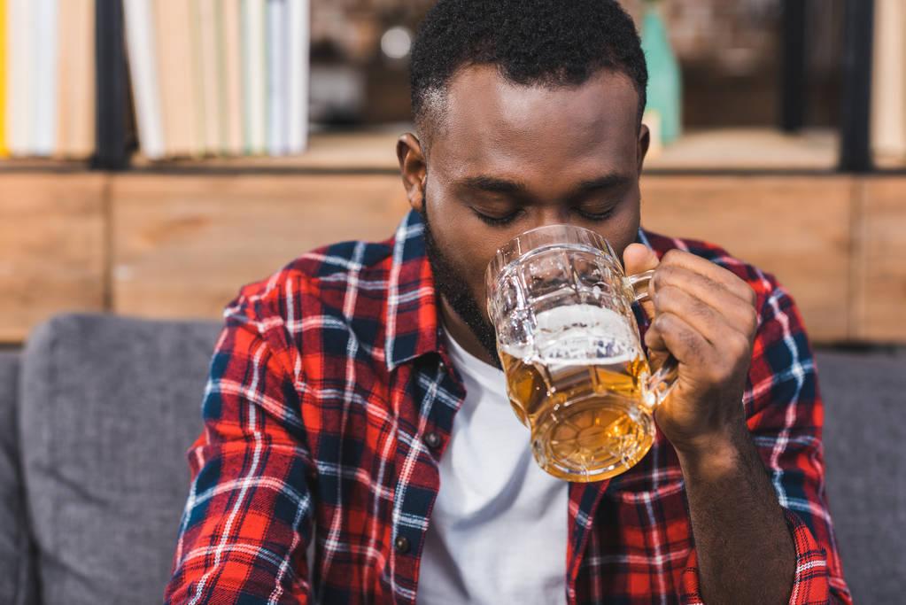 jonge Afro-Amerikaanse man bier drinken zittend op de Bank thuis - Foto, afbeelding