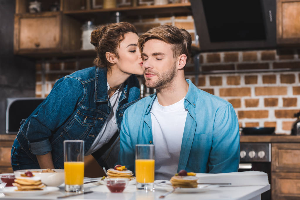 Mädchen küsst Freund am Tisch mit Saft in Gläsern und Pfannkuchen - Foto, Bild
