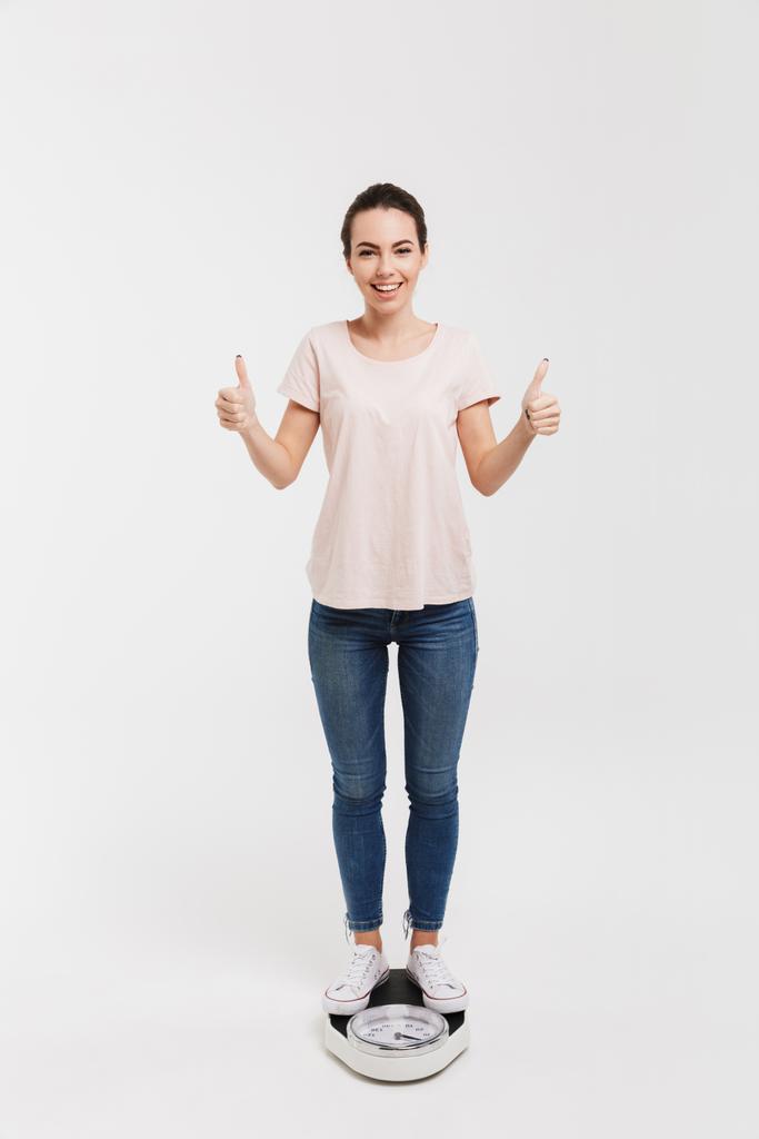 jovem mulher mostrando polegares para cima enquanto em pé em escalas isoladas em branco
 - Foto, Imagem