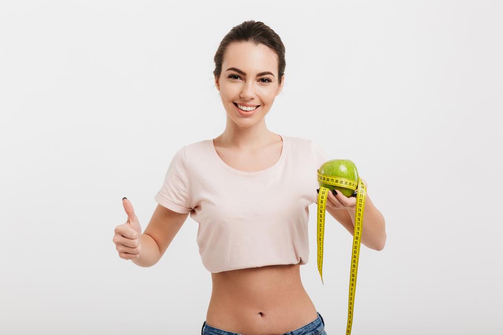 mujer sosteniendo manzana atada con cinta métrica y mostrando el pulgar hacia arriba aislado en blanco
 - Foto, imagen