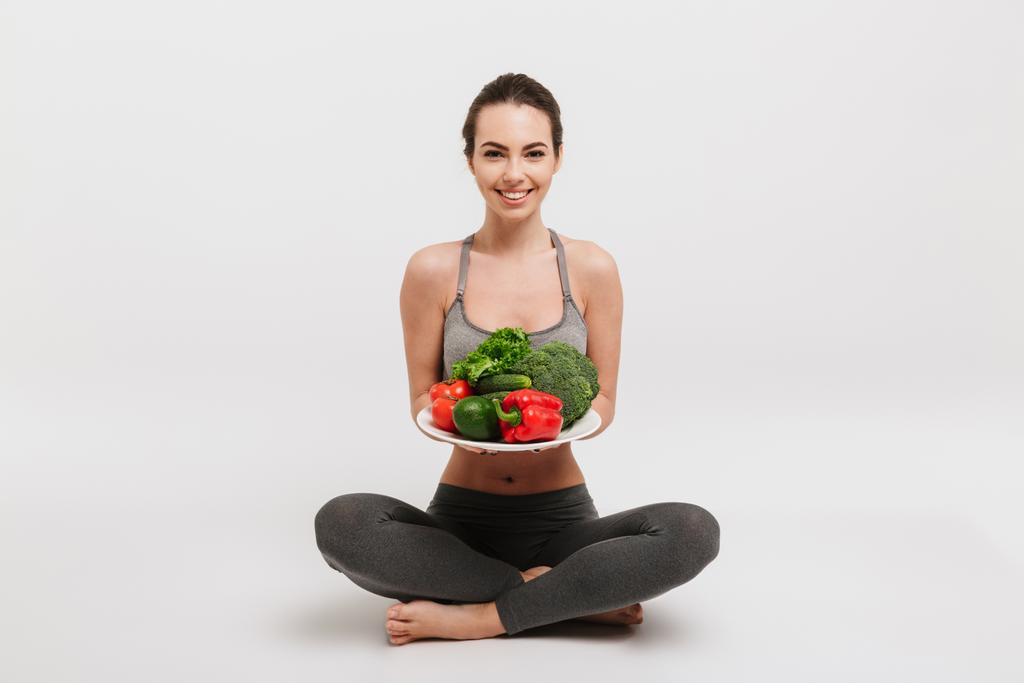 bela jovem mulher sentada no chão com bandeja de vários vegetais saudáveis isolados em branco
 - Foto, Imagem